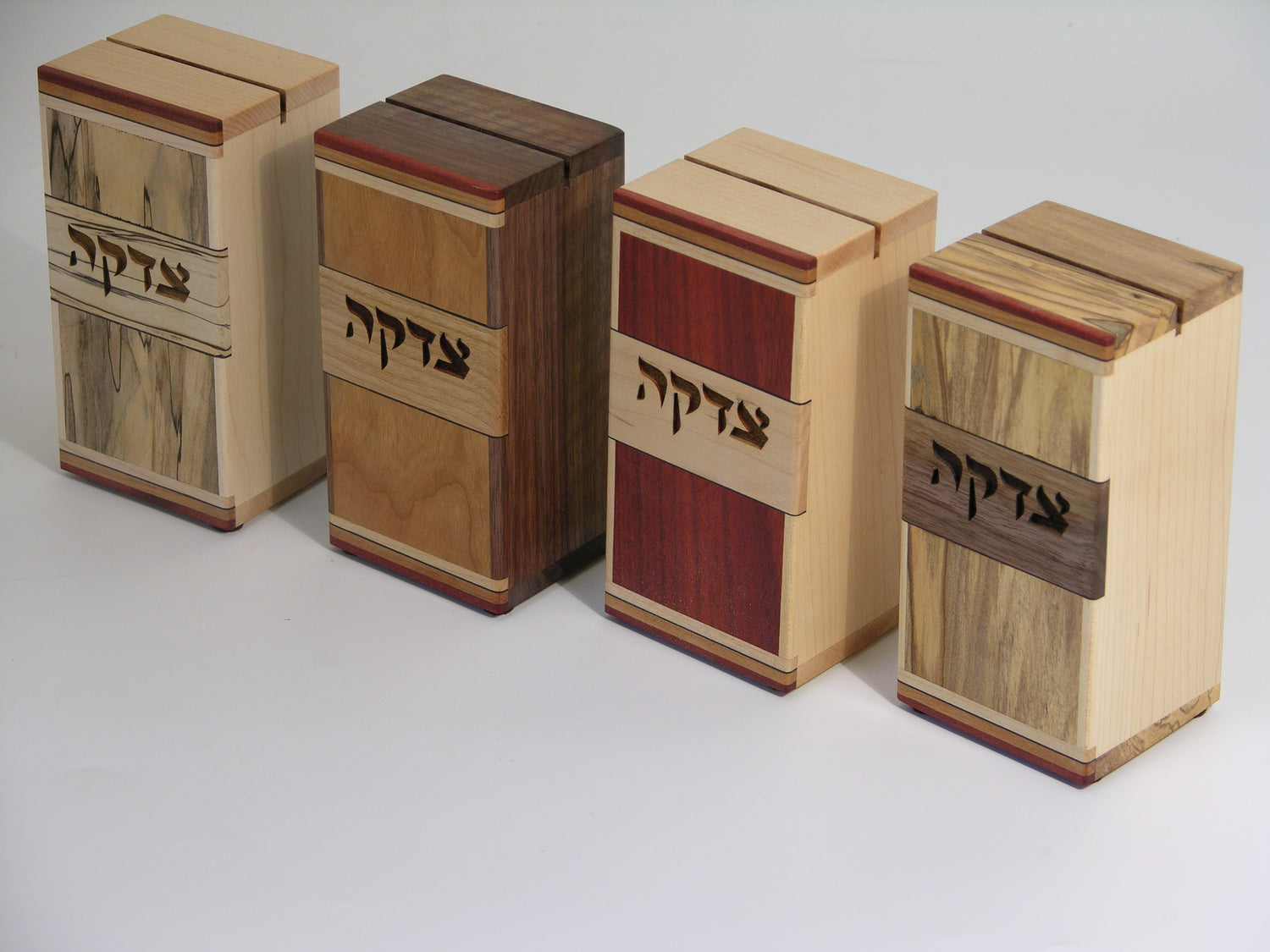 Set of four EC11 tzedakah boxes, left to right: EC11A spalted maple/maple, EC11B cherry/walnut, EC11C bloodwood/maple, EC11D ambrosia maple/maple; inlaid hardwood strips at top and bottom.